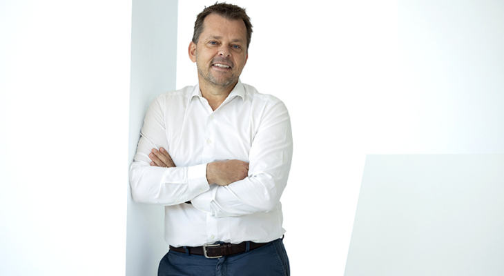 A professional portrait of Martin Mayer showing him smiling confidently while wearing a white shirt. The background is softly lit and neutral, emphasizing his face and friendly expression.