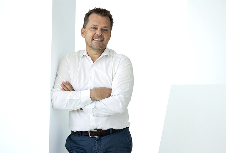A professional portrait of Martin Mayer showing him smiling confidently while wearing a white shirt. The background is softly lit and neutral, emphasizing his face and friendly expression.