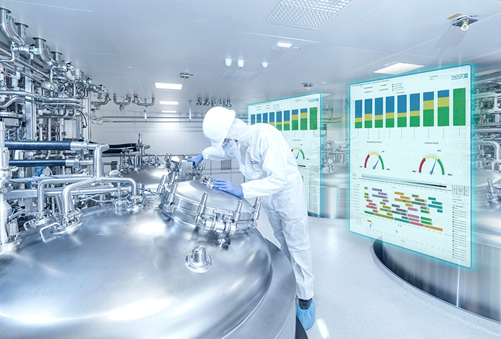 A person in protective clothing working with large metal equipment in a clean industrial setting. There are two transparent screens showing charts and graphs, suggesting the use of advanced technology to monitor processes.