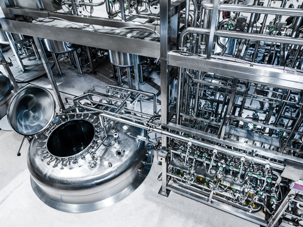 The image shows a polished, stainless steel industrial setup with a large open-top cylindrical tank surrounded by pipes and valves. It’s part of a clean, high-precision processing facility.