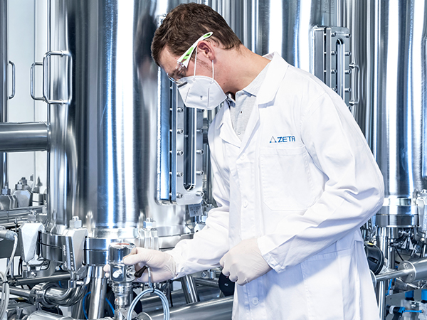 Person with lab coat, face mask and protective gloves handling a device in front of a manufacturing skid