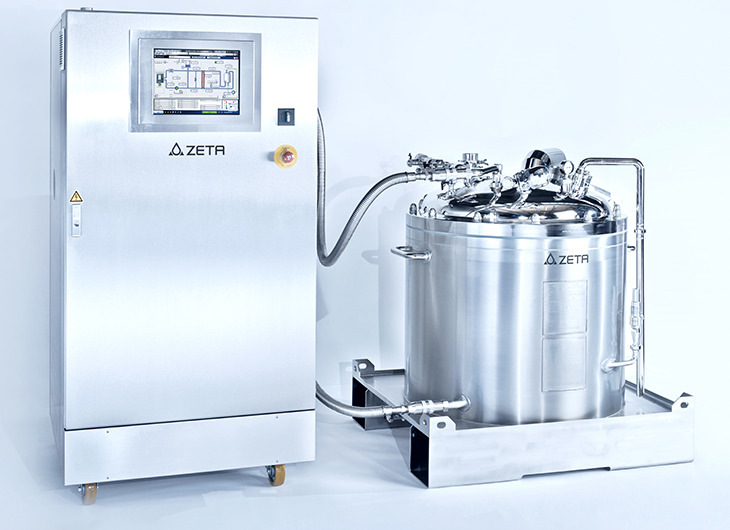 The image shows two pieces of industrial equipment made of shiny metal. One is a control unit with a screen and buttons, including an emergency stop. The other is a large tank with pipes and valves. Both are labeled “ZETA” and used in clean, precise manufacturing settings.