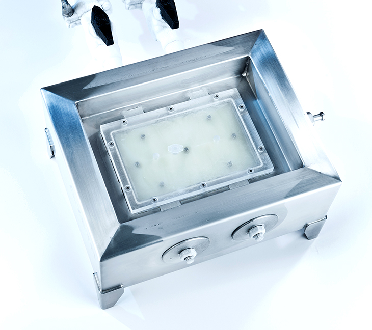 A shiny, rectangular metal device with a clear window in the middle is shown. It has two round knobs on one side and tubes connected at the top.