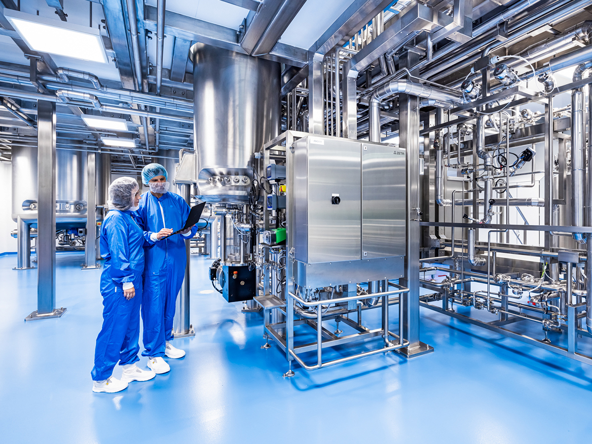 The image shows a clean industrial facility with two people in protective clothing standing on a blue floor. They are surrounded by stainless steel tanks, pipes, and control panels, likely in a pharmaceutical or food processing plant. One person holds a clipboard or tablet.