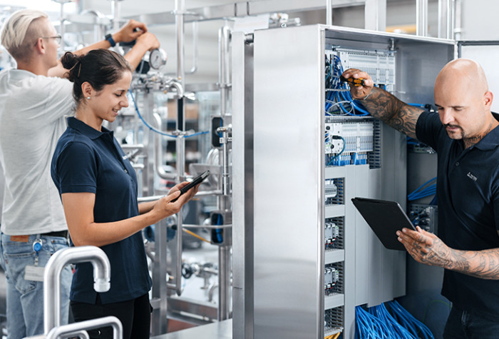 The image shows three people working together in an industrial setting. One is adjusting equipment with pipes and valves, another is using a tablet to monitor or record data, and the third is working inside an open electrical cabinet with wires, also using a tablet.