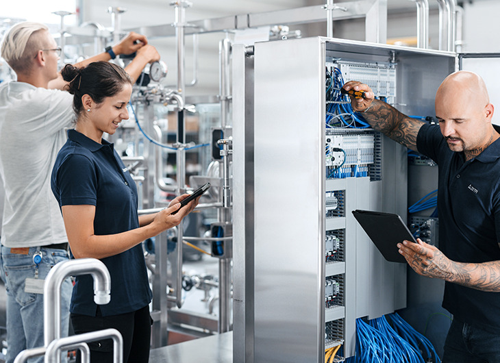 The image shows three people working together in an industrial setting. One is adjusting equipment with pipes and valves, another is using a tablet to monitor or record data, and the third is working inside an open electrical cabinet with wires, also using a tablet.