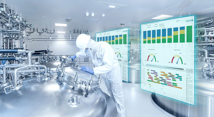 The image shows a person in protective clothing working with large metal equipment in a clean industrial setting. Behind them are two transparent screens showing charts and graphs, suggesting the use of advanced technology to monitor processes.