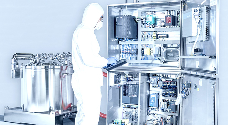 The image shows a person in protective clothing working with industrial equipment. They’re using a tablet near an open control panel full of wires and electronics. Next to them is a large metal tank with pipes and valves.