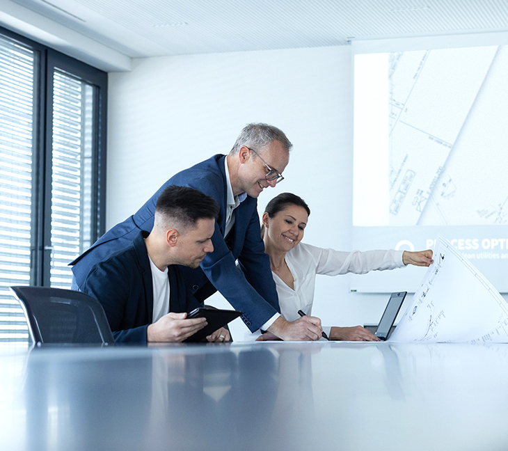Three people in a meeting room. discussing a plan which is placed on the table in front of them. One person is standing while the other two are sitting.