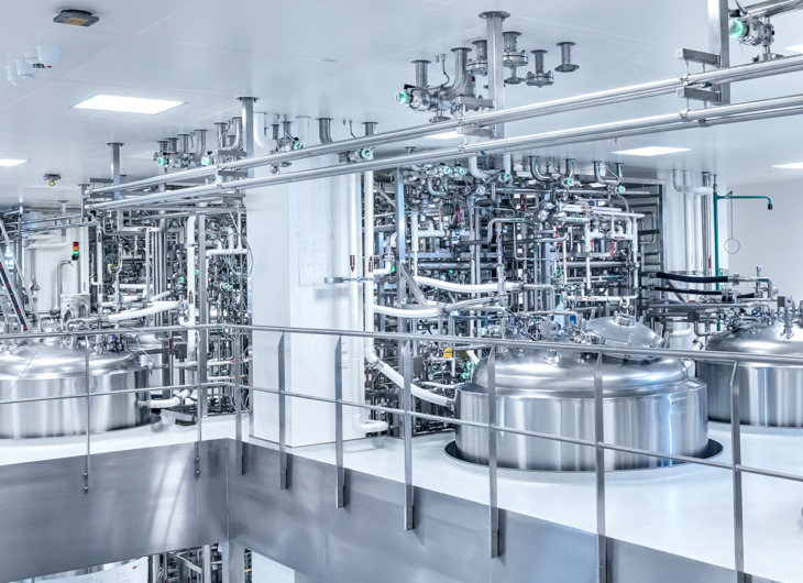 The image shows a clean, modern industrial facility with large stainless steel tanks on an elevated platform, surrounded by pipes, valves, ladders, and control panels.