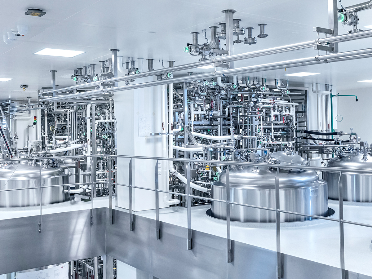 The image shows a clean, modern industrial facility with large stainless steel tanks on an elevated platform, surrounded by pipes, valves, ladders, and control panels.