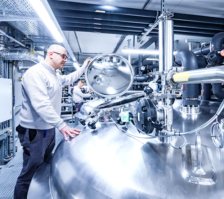 The image shows a person working with large stainless steel equipment in a clean, well-lit industrial or lab setting. The setup includes pipes, valves, and controls, likely used in pharmaceutical or food production.