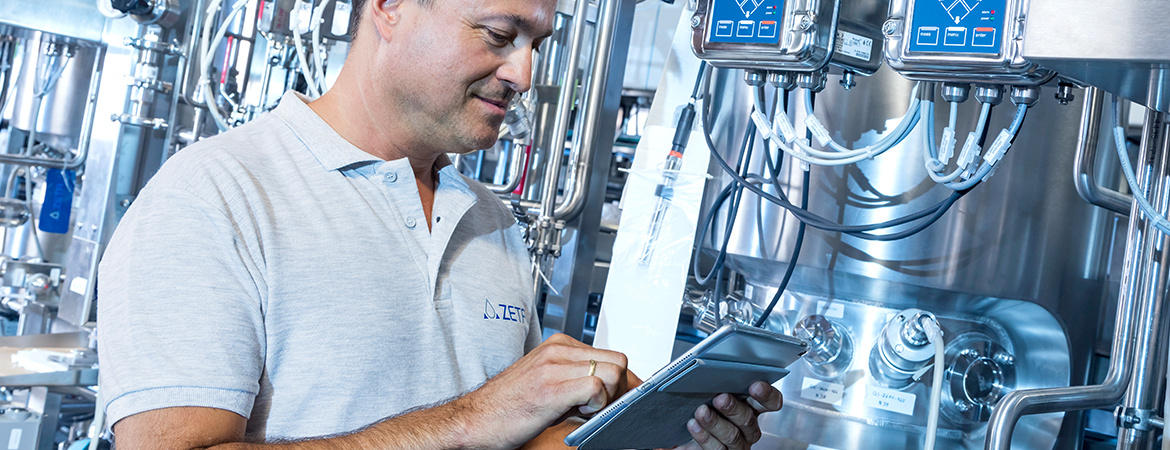 A person uses a tablet in front of stainless steel industrial machinery, likely monitoring or controlling equipment in a high-tech manufacturing setting.
