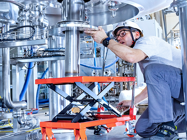 A person in safety gear uses a red hydraulic lift table to adjust a large metal component in an industrial setting with pipes and valves.