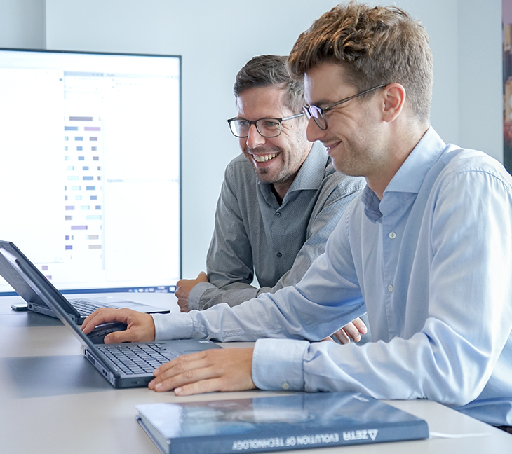 Two people working together at a desk with a laptop, reviewing data displayed on a large monitor. A book is placed on the table in the foreground.