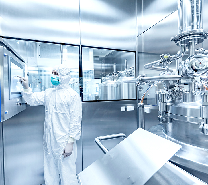 A person in full protective gear—suit, gloves, and mask—is using a touchscreen on the wall in a clean, sterile room. The space contains large stainless steel machines, likely used in pharmaceutical or biotech manufacturing.