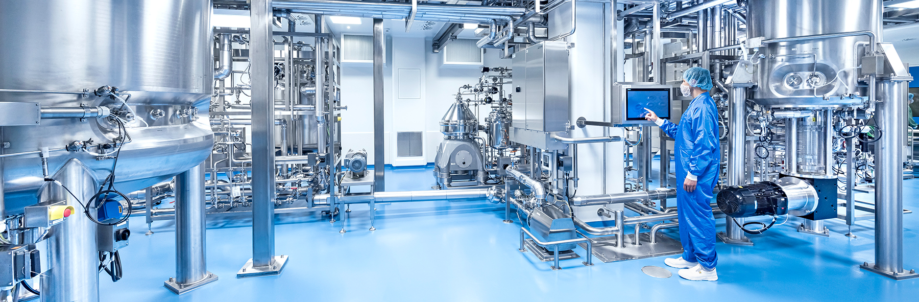 The image shows a clean, high-tech industrial facility with stainless steel tanks, pipes, and control panels. A person in protective clothing is operating a touchscreen panel.