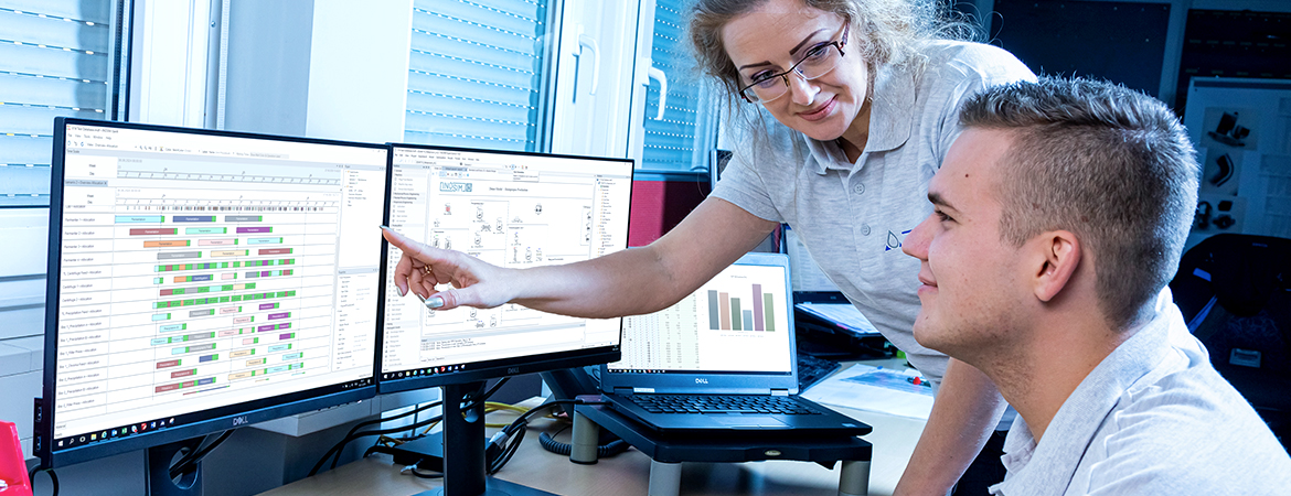 Two people working at a desk with dual computer monitors displaying scheduling charts, diagrams, and data tables. One person is seated, looking at the screens, while the other is standing and pointing at one of the monitors.
