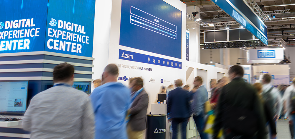 Exhibition booth at a trade fair showcasing ZETA’s Digital Experience Center with large digital displays and visitors walking through the event.