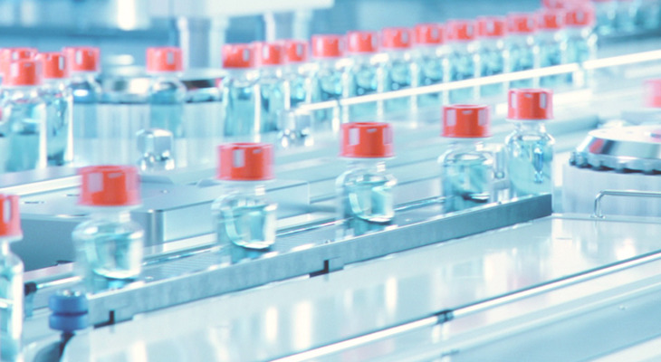 Close-up view of a pharmaceutical production line with multiple glass vials filled with a clear blue liquid, each sealed with a red cap, moving along a conveyor system in a sterile manufacturing environment.