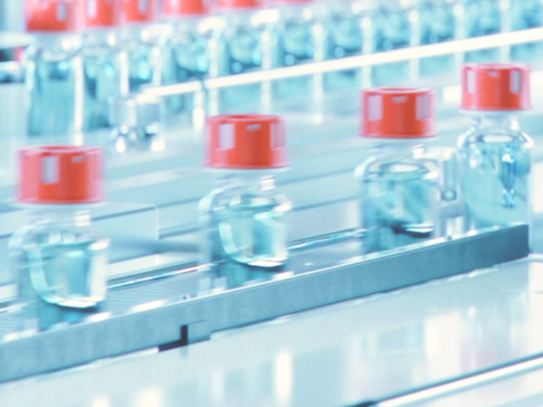 Close-up view of a pharmaceutical production line with multiple glass vials filled with a clear blue liquid, each sealed with a red cap, moving along a conveyor system in a sterile manufacturing environment.