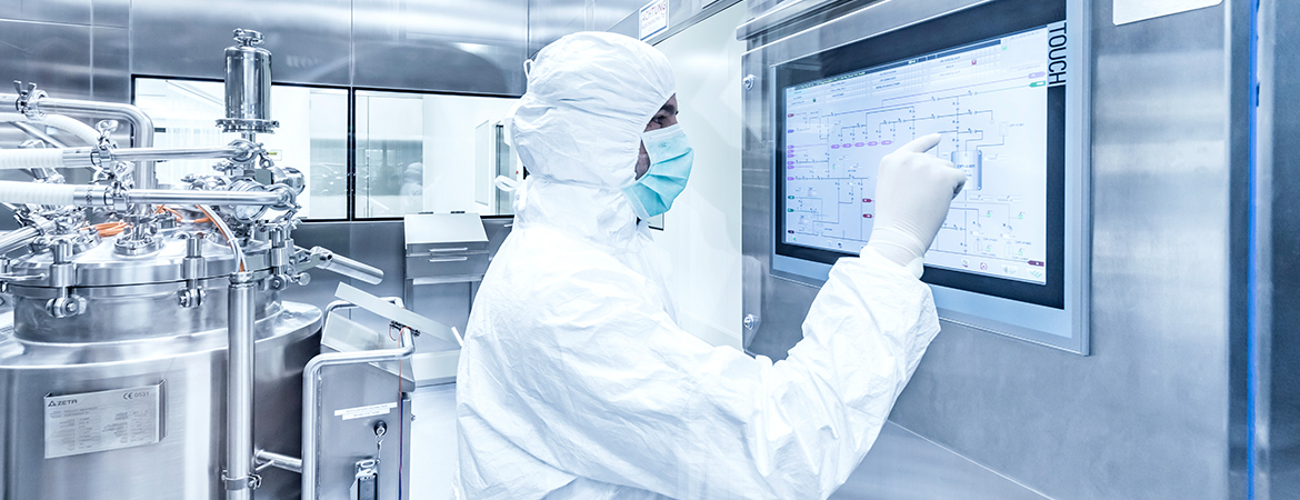 A person in full protective gear operates a touch screen in a clean, sterile lab with complex stainless steel machinery.