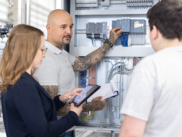 Three people are standing in front of an open electrical control panel. One holds a tablet, another a clipboard with papers, and the third has their back to the camera.