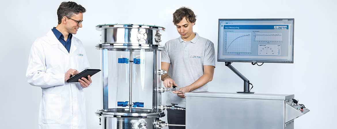 The image shows two people in a lab. One holds a tablet, while the other adjusts a large stainless steel device with transparent sections and valves. A nearby monitor displays graphs and data labeled “kLa-Measuring” with the ZETA logo.