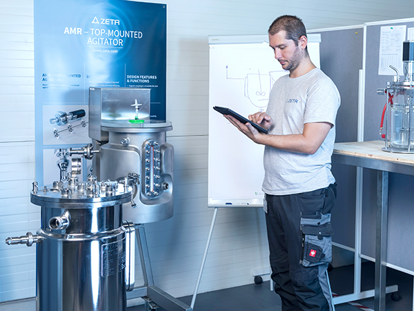 The image shows a person in a lab or industrial setting holding a tablet. Behind them is a large stainless steel machine labeled 