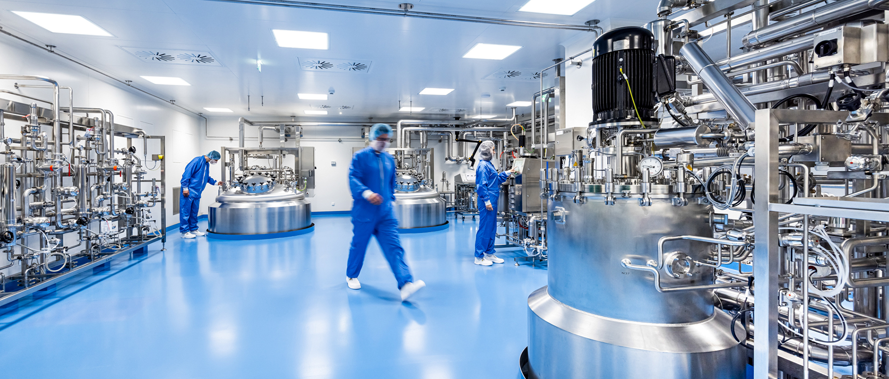 A clean, modern industrial room with shiny metal tanks, pipes, and machines. Three people in blue protective suits and face masks are working.
