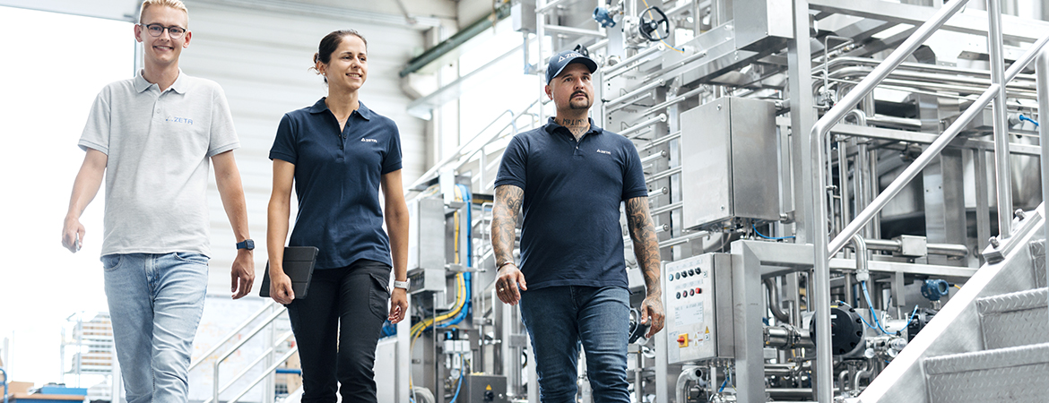 Three people in work attire walking inside a bright industrial setting, surrounded by stainless steel machinery, pipes, and control panels.