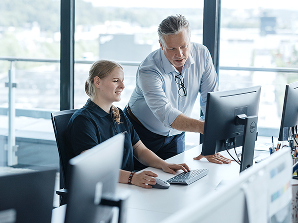 Two people in a modern office setting working together at a desk with computer monitors. One person is seated using a keyboard and mouse, while the other is standing and pointing at the screen.
