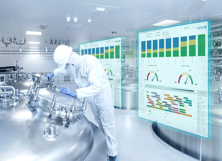 A person in protective clothing working with large metal equipment in a clean industrial setting. There are two transparent screens showing charts and graphs, suggesting the use of advanced technology to monitor processes.
