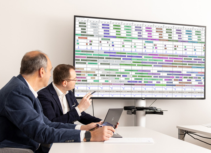 Two people seated at a conference table analyzing a large wall-mounted display showing a detailed, color-coded scheduling chart with multiple rows and timelines.